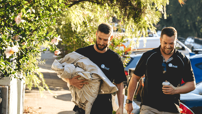 two tradies walking along the sidewalk one holding a canvas drop sheet and the other holding a cup of coffee