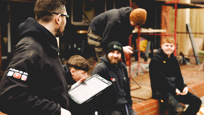 group of tradies sitting on the deck of a house building looking at another tradie holding a tablet