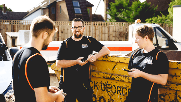 group of tradies standing outside next to skip bin holding cellphones in hand while talking